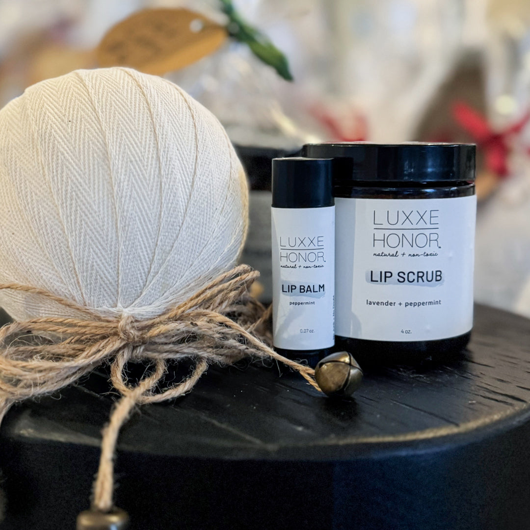 A cream-colored yarn ball sits beside a peppermint lip balm and a lavender-peppermint lip scrub on a dark surface.  The products are labeled "LUXXE HONOR, natural + non-toxic".
