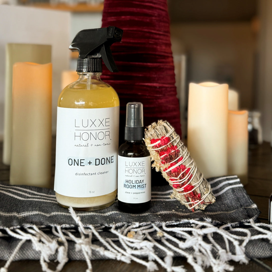 A disinfectant cleaner and room mist sit on a table, next to a smudge stick and candles.  The bottles are labeled "LUXXE HONOR," "ONE + DONE disinfectant cleaner," and "HOLIDAY ROOM MIST clove + peppermint."